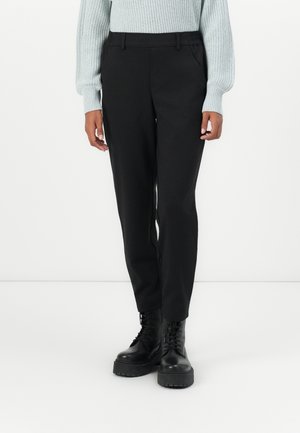 Persona que lleva pantalones negros de pierna recta, suéter de punto gris claro y botas negras gruesas con cordones, de pie contra un fondo blanco.