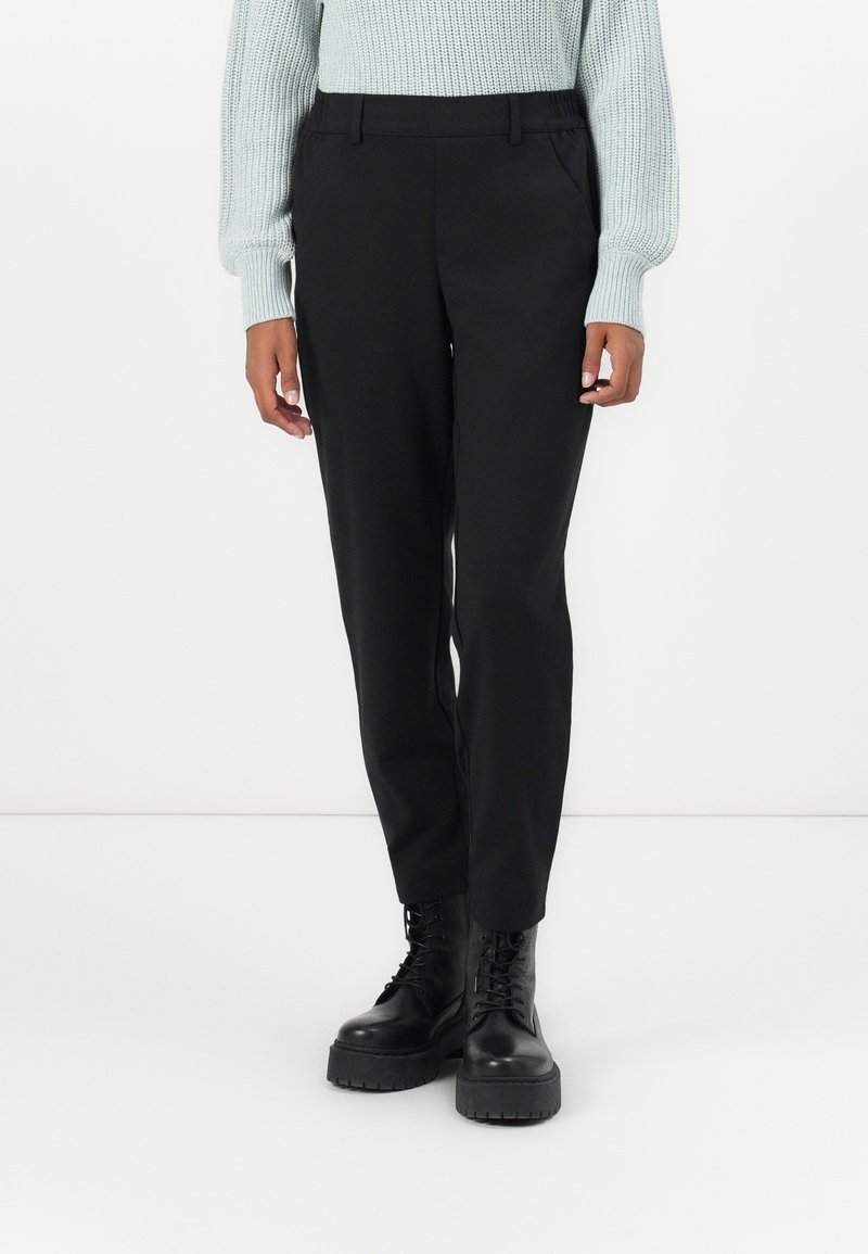 Personne portant un pantalon noir droit, un pull en maille gris clair et des bottes noires épaisses à lacets, debout sur un fond blanc.