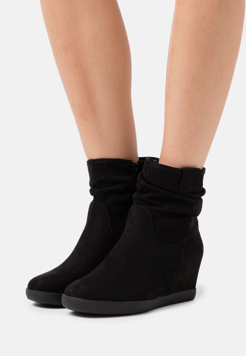 Jambes portant des bottes compensées en daim noir mi-mollet avec tige souple et fermeture éclair latérale, sur fond blanc.