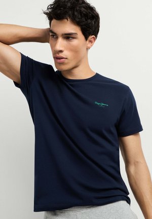 Joven con el pelo rizado y oscuro, vistiendo una camiseta azul marino de Pepe Jeans y pantalones grises, posando con una mano detrás de la cabeza.