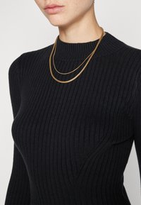 Črna rebrasta pulover s visokim ovratnikom, ki ima teksturirano površino. Ovratnik krasi plastičen zlatih metalnih verižic.