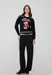 Schwarzer Strickweihnachtspullover mit einem Teddybär in einem Weihnachtsmannkostüm, mit dem Text "Frohe Weihnachten" und einem gemusterten Saum. Kombiniert mit schwarzen Hosen.