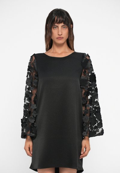Robe noire avec des manches transparentes ornées de fleurs. Texture douce, silhouette simple, encolure ronde et longueur au genou.