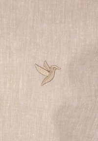 Beez kangas, millel on tekstureeritud pind ja väikese beeži tikandiga kolibri kujundus.