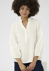 Blusa bianca in lino con colletto a volant, chiusura a bottoni e maniche a sbuffo. Texture morbida, vestibilità rilassata e un design semplice ed elegante.