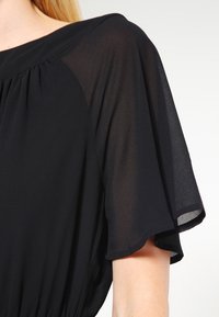 Tissu noir transparent, manches courtes flottantes, décolleté drapé, avec une texture subtile et des détails doucement froncés autour des épaules.