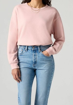Femme portant un sweat-shirt rose clair à manches longues rentré dans un jean taille haute bleu clair, avec une main dans la poche et des colliers en or superposés.