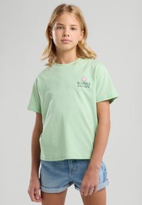 T-shirt in cotone verde chiaro con maniche corte, con una grafica a conchiglia e il testo "SUMMER ESCAPE" in verde scuro. Indossato con pantaloni corti in denim.