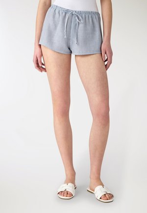 Donna che indossa pantaloncini a righe bianche e azzurre con coulisse e sandali bianchi a fascia, in piedi davanti a uno sfondo chiaro e uniforme.