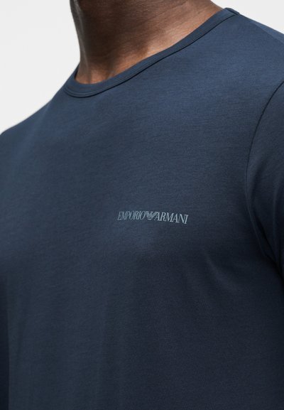 Άνδρας που φοράει μπλε ναυτικό t-shirt με στρογγυλή λαιμόκοψη και μικρό ανοιχτό γκρι λογότυπο Emporio Armani στο αριστερό στήθος.