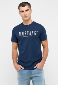 Tumši zila kokvilnas T-kreklu ar apaļu kakla izgriezumu un īsām piedurknēm, ar baltu "MUSTANG" logo, kas izceltas uz krūtīm.