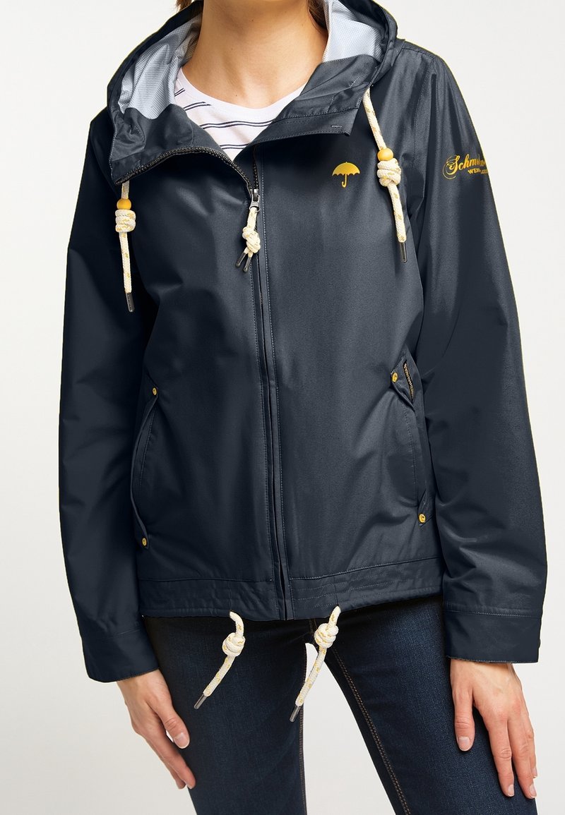 Marinblå vattentät jacka med huva, med gul paraplylogotyp, dragsko och detaljer i guldton. Texturerat tyg, blixtlåsförsedda fickor.