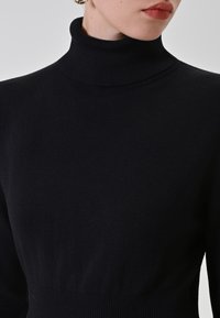 Donna che indossa un maglione a collo alto nero e orecchini a cerchio dorati, mostrata dalle spalle alle labbra su uno sfondo semplice.