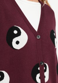 Burgundijas kardigāns ar yin-yang rakstu melnbaltā krāsā. Ar V-veida kaklu un diviem melniem pogām priekšā. Mīksta tekstūra.