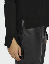 Maglione nero lavorato a maglia con orlo a costine e spacchi laterali, abbinato a eleganti pantaloni in pelle nera. Primo piano di una mano appoggiata sui pantaloni.