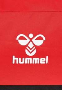 Bolsa de tela roja con un logo blanco que presenta un diseño abstracto de una abeja sobre la palabra "hummel" en letras grandes y claras.
