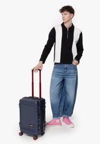 Hårdkantad resväska i marinblått med röda handtag och hjul. Modellen bär en svart och vit zip-tröja, löst sittande blå jeans och rosa sneakers.