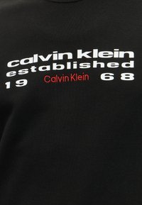 Μαύρο ύφασμα με λευκό κείμενο «calvin klein established 1968» και μικρότερο κόκκινο «Calvin Klein» τοποθετημένο στο κέντρο παρακάτω.