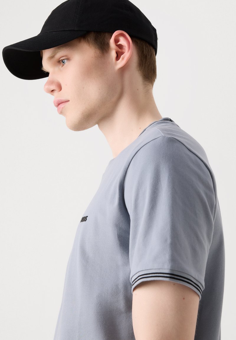 T-shirt en coton gris avec logo noir, présentant des manches courtes et une casquette de baseball noire, vue de côté. Les détails incluent un poignet rayé.