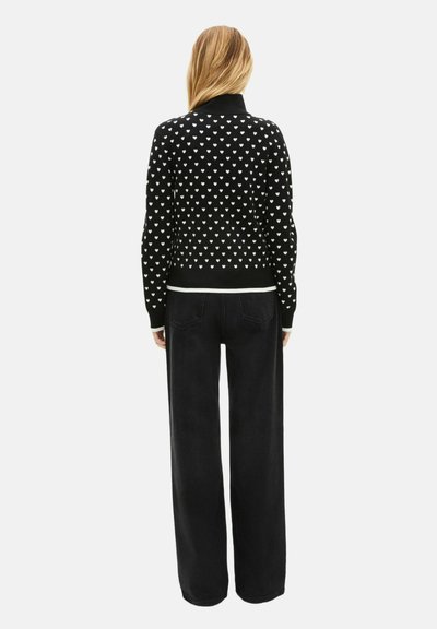 Cardigan noir avec motif de cœurs blancs, poignets et ourlet côtelés, associé à un pantalon en denim foncé taille haute. La texture semble douce et tricotée.