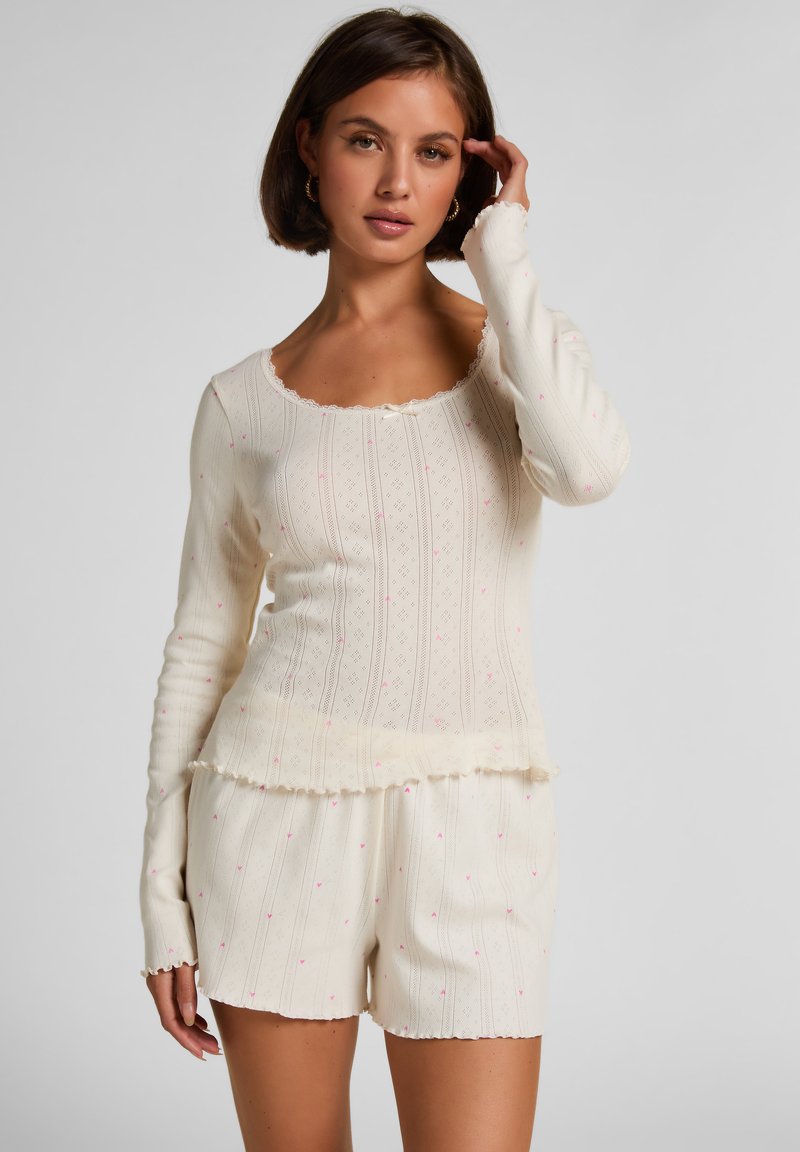 Μπλούζα μακριμάνικη χρώματος κρέμα με σκαλιδωτή λαιμόκοψη και υφασματινό σχέδιο· συνδυασμένη με κοντά παν τελονάκια στο ίδιο χρώμα με ροζ λεπτομέρειες.