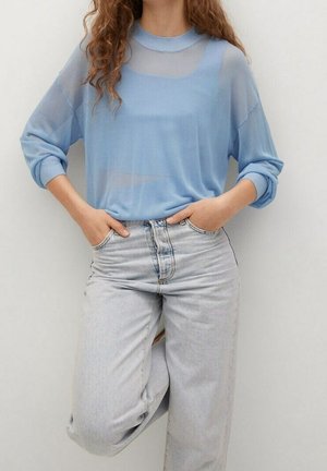 Kvinde iført en gennemsigtig lys blå langærmet top med synlig undertrøje, kombineret med højttaljede lysegrå jeans, hænderne i lommerne.