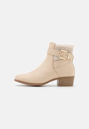 Bottine beige à bout arrondi, doublure en fausse fourrure, accent de boucle dorée et talon empilé avec des rayures horizontales. Matériau suédé.
