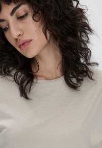 Hellbeige Rundhalsausschnitt-Oberteil aus weichem Stoff. Besitzt kurze Ärmel und eine subtile Textur. Das Haar ist dunkel und lockig, teilweise das Gesicht umrahmend.