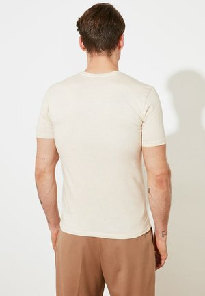 Mann mit kurzen braunen Haaren, der ein schlichtes off-white kurzärmeliges T-Shirt und hellbraune Hosen trägt, von hinten vor einer weißen Wand gezeigt.
