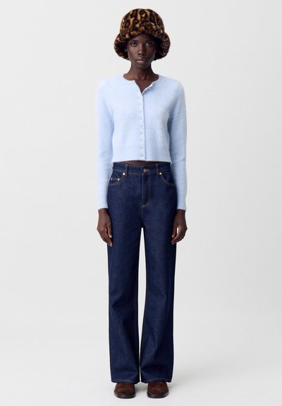 Cardigan corto azzurro chiaro con bottoni frontali, abbinato a jeans a vita alta in denim scuro e un cappello maculato marrone.