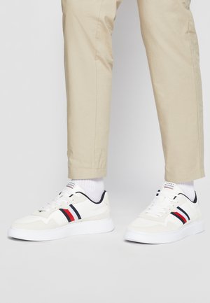 Sneaker athletic bianche con strisce rosse e blu navy, parte superiore in suede testurizzato e mesh, suola bianca, abbinate a pantaloni beige e calze bianche.