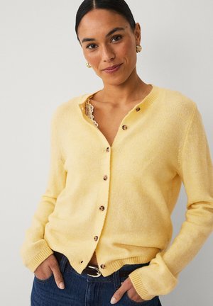 Vrouw met donker haar draagt een gele cardigan met knopen over een top met kanten rand, blauwe spijkerbroek, gouden oorbellen, staand tegen een effen achtergrond.