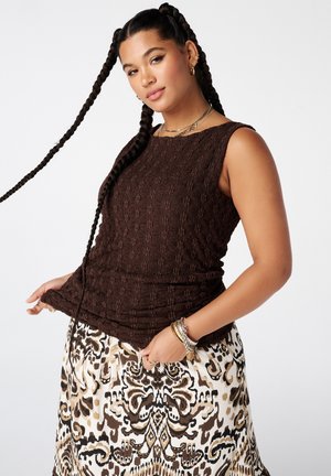 Femme aux longues tresses portant un haut texturé marron sans manches et une jupe à motifs, accessoirisée de bracelets et de colliers, sur un fond uni.