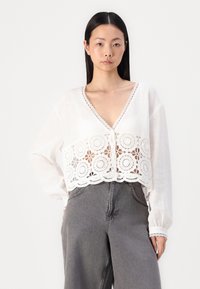Blouse blanche courte à manches longues, ornée de détails en dentelle délicate et d'un col en V. Associée à un pantalon large gris.