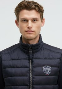 Gilet puffo blu marina con colletto alto, chiusura con zip e toppa con logo sul petto. Presenta una texture trapuntata e lati in tessuto a contrasto.