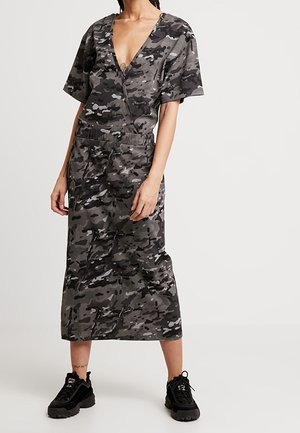 Robe à motif camouflage dans des tons de gris avec un décolleté en V profond, des manches courtes, une taille élastique et une longueur jusqu'aux chevilles, accompagnée de baskets noires.