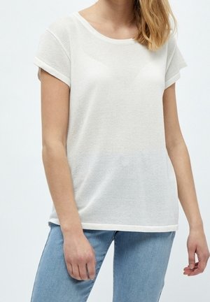 Kobieta ubrana w białą teksturowaną koszulkę z krótkim rękawem w połączeniu z jasnoniebieskimi dżinsami, widziana od szyi do bioder na jednolitym tle.