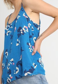 Donna che indossa un top a maniche corte blu con fiori e jeans di denim blu chiaro, con la mano appoggiata sui fianchi e un tatuaggio visibile sull'avambraccio.