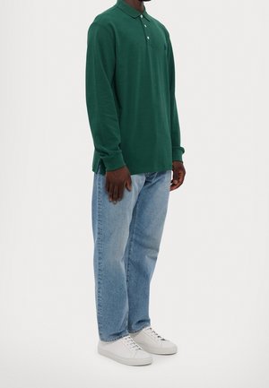 Mann trägt ein dunkelgrünes Polohemd mit langen Ärmeln, hellblaue Jeans und weiße Turnschuhe, steht vor einem schlichten weißen Hintergrund.