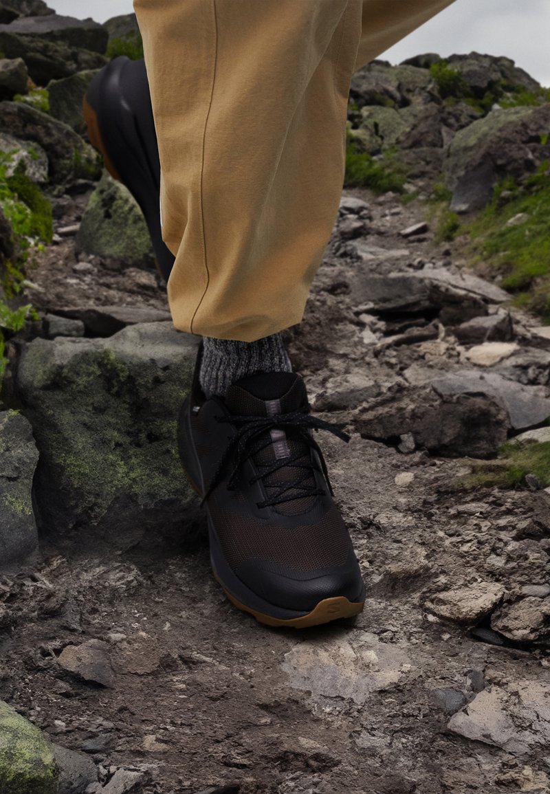 Schwarzer Wanderschuh mit strukturiertem Mesh-Obermaterial und Gummisohle, kombiniert mit hellbeigen Hosen und grauen Socken, auf felsigem Gelände.