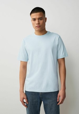 Junger Mann mit kurzen Haaren, der ein hellblaues kurzärmeliges T-Shirt und blaue Jeans trägt, steht vor einem schlichten hellen Hintergrund.