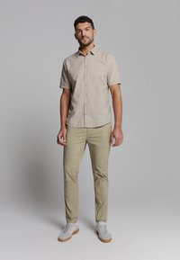 Korte mouwen beige overhemd met knopen, lichte kaki broek, grijze sneakers met witte strepen, staande houding, neutrale achtergrond.