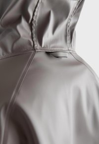 Giacca impermeabile grigia chiara con texture liscia, caratterizzata da un alto colletto e un piccolo anello di attacco vicino alla cucitura posteriore.