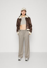 Hnedá kožená bunda s krémovým kožušinovým golierom, šedý crop top, široké nohavice s hnedo-bežovým károvaným vzorom, béžová čiapka a strieborné tenisky.