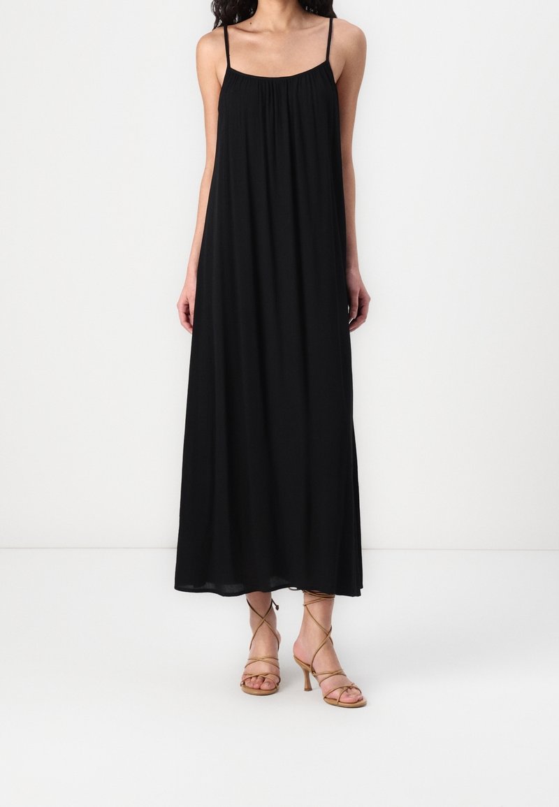 Černé maxi šaty s tenkými ramínky, lehkým materiálem, řaseným výstřihem a splývavou siluetou. V kombinaci s béžovými sandály na podpatku.