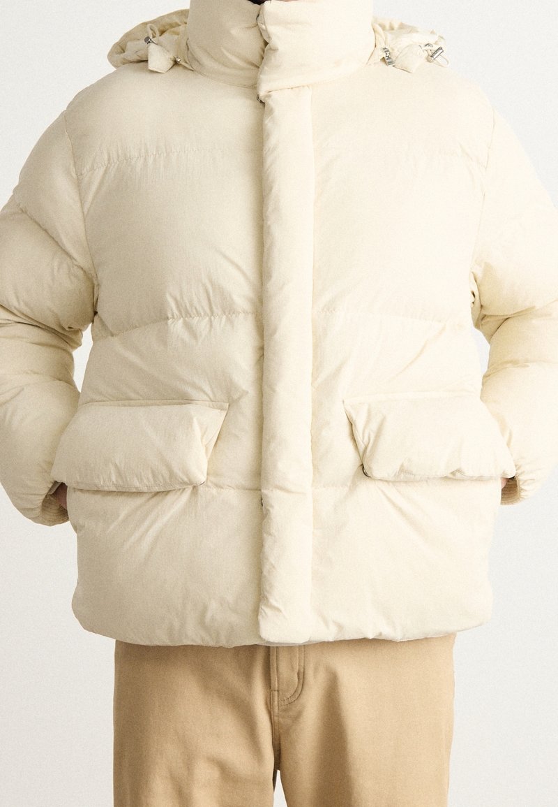 Beige pufferjakke med høj krave, to store frontlommer og en glat tekstur. Designet med en skjult frontlynlås og trykknapper.