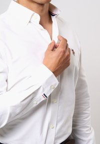 Valkoinen kauluspaita, jossa napillinen kaulus, rintakehän kirjailulla ja raitakuvioilla kalvosimissa.