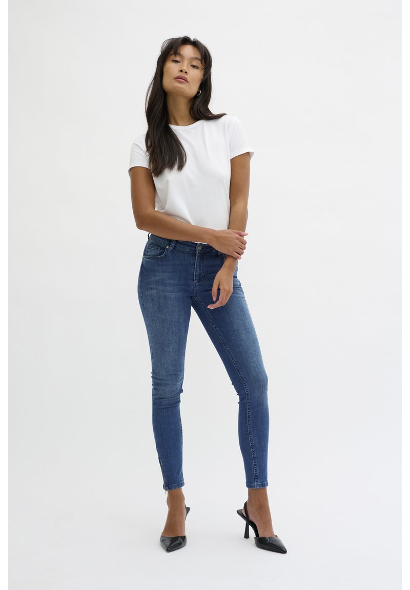 Fehér rövid ujjú póló sötétkék szűk farmernadrággal és fekete magassarkú cipővel párosítva. Egyszerű dizájn, sima anyag és testhezálló forma.