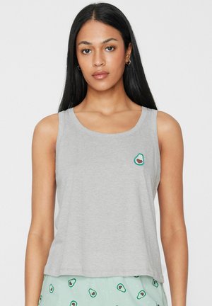 Femme portant un haut sans manches gris clair avec un petit motif avocat et un pantalon vert menthe assorti avec motif avocat, debout sur fond blanc.