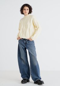 Pull en maille crème à col montant, associé à un jean bleu large et des bottes noires à semelles épaisses, sur fond blanc.